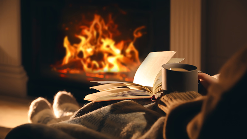 Person sitting on a sofa in front of a fireplace, reading and drinking a warm drink