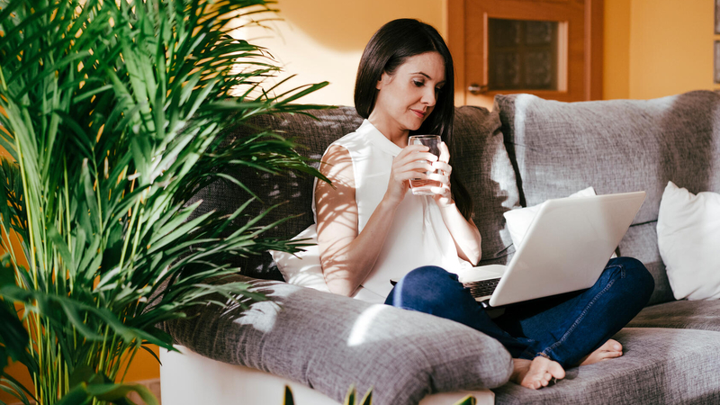 Woman sitting on the sofa drinking water while working on her laptop computer