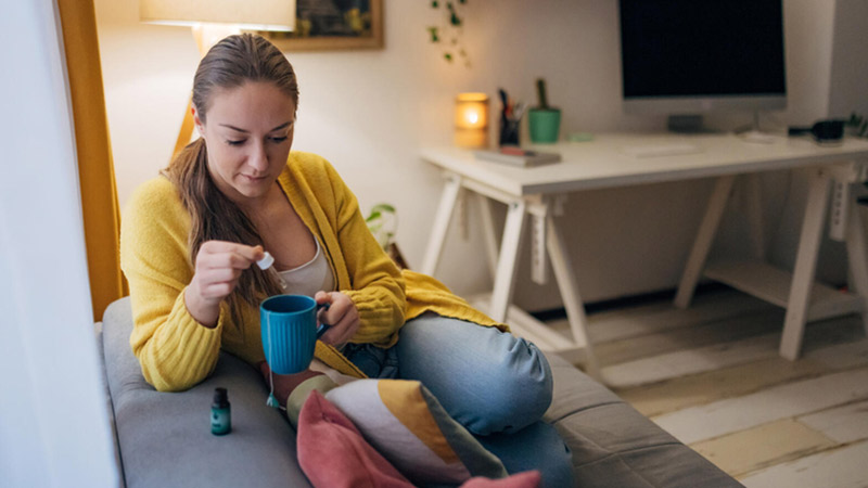 Woman sitting on a sofa adding CBD extract to a cup of tea