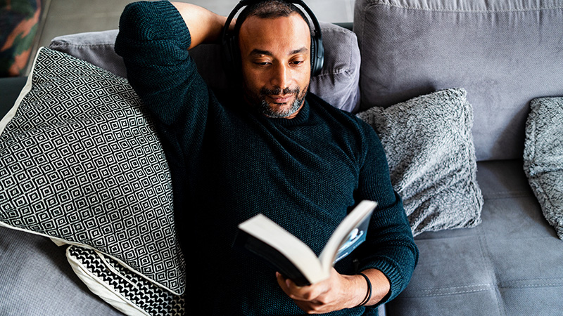 Man reclining on a sofa wearing headphones and reading a book